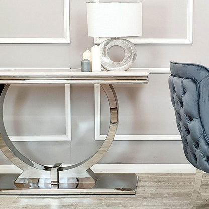 Elegant Arriana Console table by Furnish 365 Limited in a stylish hallway, featuring a sleek glass top and polished stainless steel circular base alongside decorative elements like a lamp and candles.