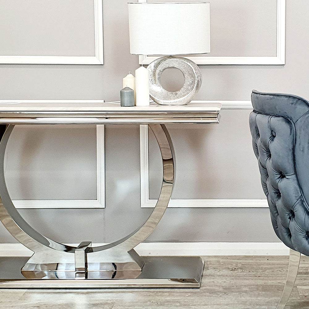 Elegant Arriana Console table by Furnish 365 Limited in a stylish hallway, featuring a sleek glass top and polished stainless steel circular base alongside decorative elements like a lamp and candles.